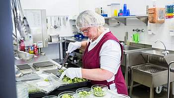 Eine Frau mit roter Schürze steht in einer Küche und füllt Gurkensalat ab