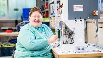 Eine Frau mit türkisem Pulli sitzt an einer Maschine und hält ein kabel in der Hand